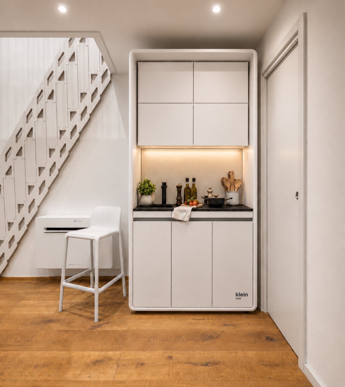 La cucina KleinCASA accanto alla scala: mobile bianco compatto con piano scuro, pensili e uno sgabello bianco