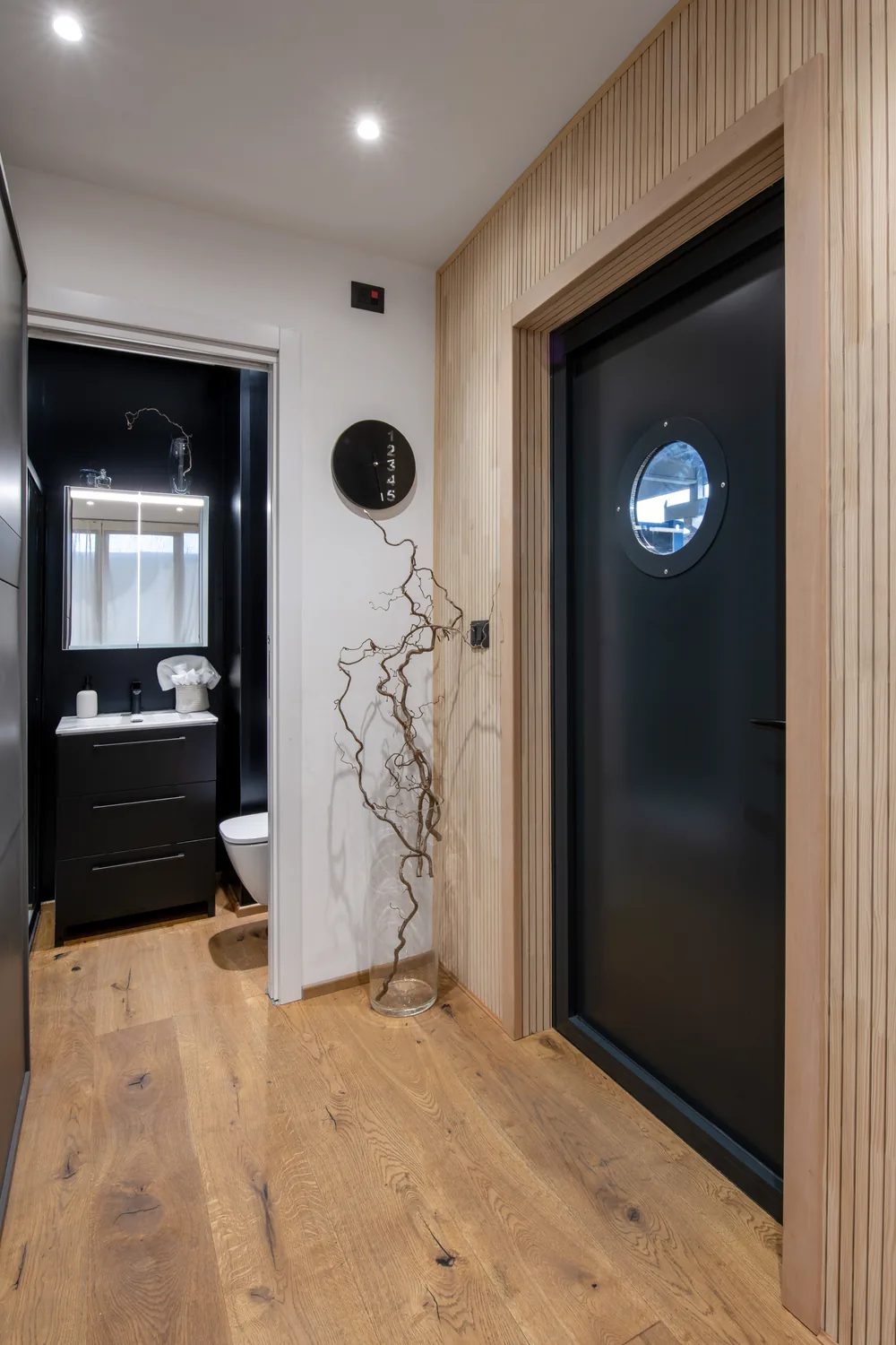 Entrata con porta scura e oblò, parete in doghe di legno e, a sinistra, il bagno