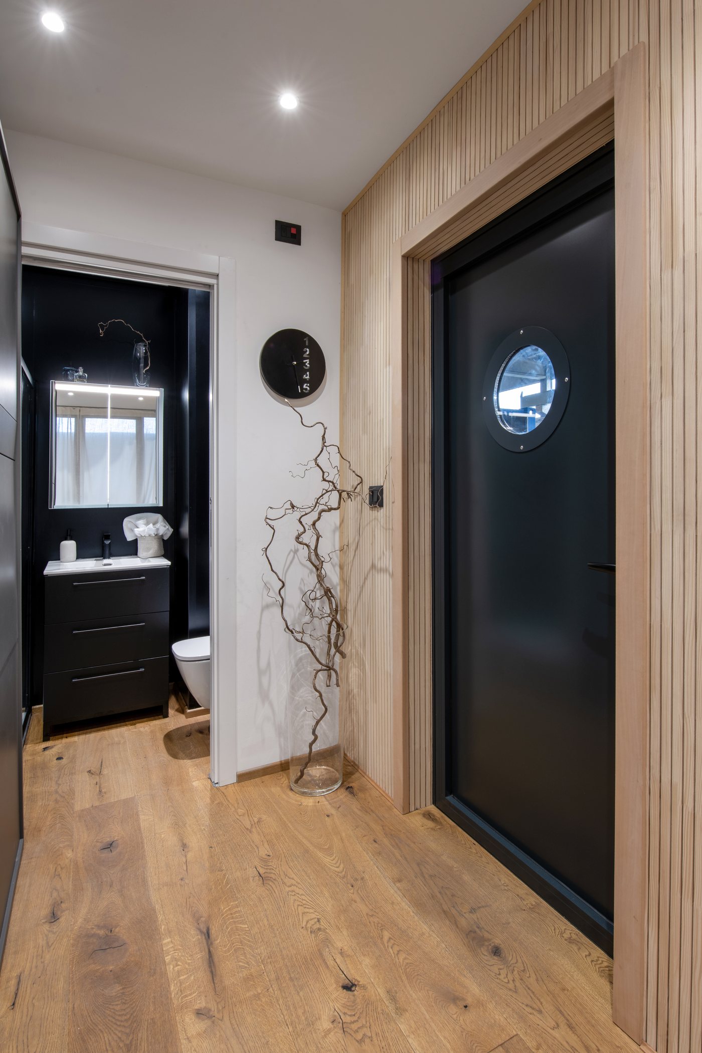 Entrata con porta scura e oblò, parete in doghe di legno e, a sinistra, il bagno